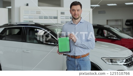 Happy man in a business suit, a manager at a car dealership holds a TabletPC with green screen in his hands and poses near the cars. Happy man in a business suit, a manager at a car dealership holds a TabletPC with green screen in his hands and poses near the cars. 125510766