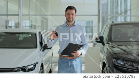 Handsome man manager at a car dealership in a business suit holding clipboard documents and car keys in his hands stands near the cars. Concept of car shopping, buying, and dealership. Handsome man manager at a car dealership in a business suit holding clipboard documents and car keys in his hands stands near the cars. Concept of car shopping, buying, and dealership. 125510792