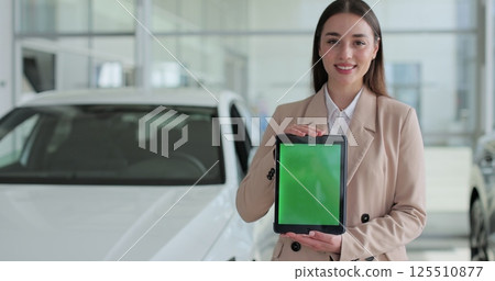 Happy woman in a business suit, a manager at a car dealership holds a TabletPC with green screen in her hands and poses near the cars. Happy woman in a business suit, a manager at a car dealership holds a TabletPC with green screen in her hands and poses near the cars. 125510877