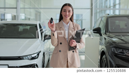 Happy woman manager at a car dealership in a business suit holding clipboard documents and car keys in her hands stands near the cars. Concept of car shopping, buying, and dealership. Happy woman manager at a car dealership in a business suit holding clipboard documents and car keys in her hands stands near the cars. Concept of car shopping, buying, and dealership. 125510886