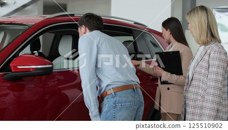 Car saleswoman showing to young couple new automobile at dealership salon. Concept of car shopping, buying, and dealership. Car saleswoman showing to young couple new automobile at dealership salon. Concept of car shopping, buying, and dealership. 125510902