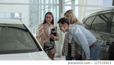 Happy family couple talking to car dealer in dealership discussing automobiles looking at new model. Concept of car shopping, buying, and dealership. 125510923