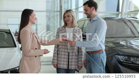 Young couple buying new car in car dealership. Saleswoman giving car keys to new car happy couple family buyers. Auto business concept. 125510955