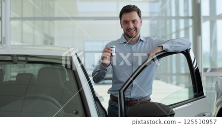 Happy man standing at dealership near new car. Handsome young male choosing transport to buy. Concept of car shopping, buying, and dealership. 125510957