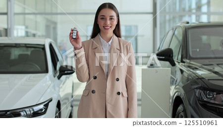 Woman manager standing near modern cars and showing keys to camera. Sales manager looking directly. Concept of car shopping, buying, and dealership. 125510961