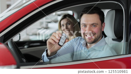 Young man showing key to new car. Happy couple family buyers clients. Auto business concept. Man holding car keys at the dealership salon. 125510981