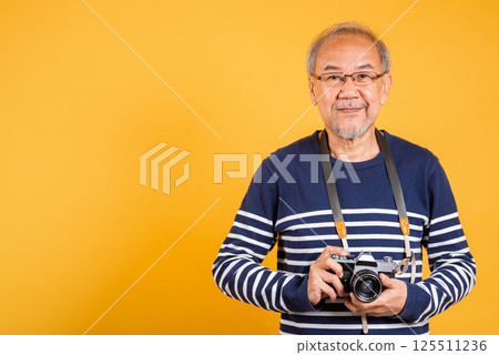 Portrait Asian old man wearing glasses holding vintage SLR camera studio shot isolated yellow background, smiling happy Photographer elderly man gray haired holds old fashioned camera Portrait Asian old man wearing glasses holding vintage SLR camera studio shot isolated yellow background, smiling happy Photographer elderly man gray haired holds old fashioned camera 125511236