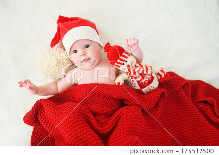 A cute baby in a Santa hat with a big bubo covered with a red knitted blanket hugging a snowman toy on a white background. 125512500