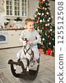 a child in gray pajamas rides a rocking horse against the backdrop of a Christmas tree and a modern kitchen. 125512508