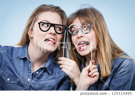 Two happy women holding fake eyeglasses on stick Two happy women holding fake eyeglasses on stick 125513495