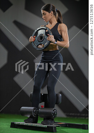 woman utilizes weight plate while performing strength training exercises in modern gym environment. focus is on her determination and effort enhance physical fitness. 125513656