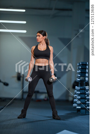 fit woman stands confidently in contemporary gym holding dumbbells in each hand. She is engaged in strength training exercise focusing on her form. fit woman stands confidently in contemporary gym holding dumbbells in each hand. She is engaged in strength training exercise focusing on her form. 125513686