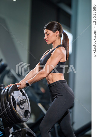 determined woman stands in fitness center preparing lift heavy weights. She wears fitted workout outfit showcasing her toned physique while concentrating on her exercise routine. determined woman stands in fitness center preparing lift heavy weights. She wears fitted workout outfit showcasing her toned physique while concentrating on her exercise routine. 125513690
