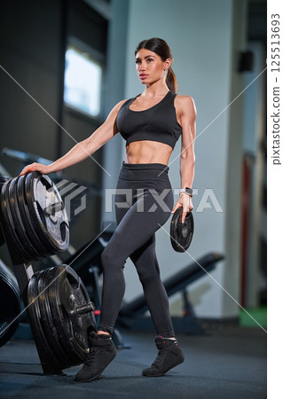 In well equipped gym fit woman performs strength training lifting weights. She wears athletic gear and demonstrates focus while working out. 125513693