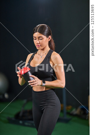 young woman stands in gym taking moment hydrate shaker cup in hand. Dressed in black athletic outfit she maintains focus amidst her workout routine. young woman stands in gym taking moment hydrate shaker cup in hand. Dressed in black athletic outfit she maintains focus amidst her workout routine. 125513695