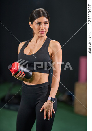 woman dressed in black athletic attire stands confidently in fitness studio while holding shaker bottle. 125513697