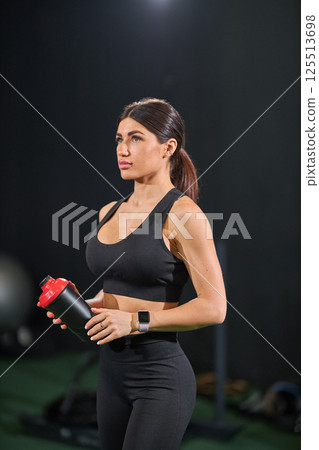 focused individual stands in fitness studio holding shaker bottle. Dressed in activewear she prepares for evening workout while surrounded workout equipment. focused individual stands in fitness studio holding shaker bottle. Dressed in activewear she prepares for evening workout while surrounded workout equipment. 125513698