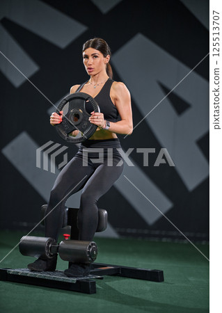 woman in activewear engages in strength training exercise gym. She sits on bench while holding weight plate demonstrating focus on her workout routine. 125513707