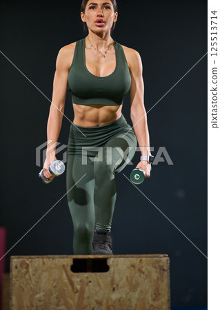 woman engages in high intensity workout routine using dumbbells while stepping onto wooden platform. She exhibits strength and focus in modern fitness studio. woman engages in high intensity workout routine using dumbbells while stepping onto wooden platform. She exhibits strength and focus in modern fitness studio. 125513714