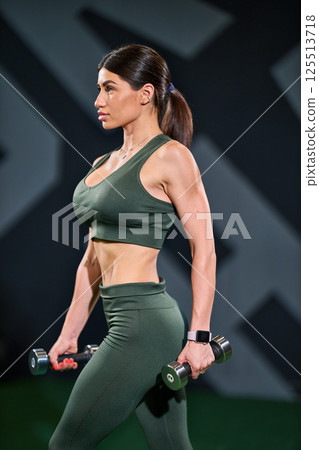 woman in athletic wear is holding dumbbells fitness studio. She demonstrates determination and strength while targeting her fitness goals. background features motivational designs. woman in athletic wear is holding dumbbells fitness studio. She demonstrates determination and strength while targeting her fitness goals. background features motivational designs. 125513718