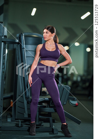 woman stands confidently in gym wearing purple sports bra and matching leggings. She showcases strength and poise next workout equipment in well lit space. 125513729