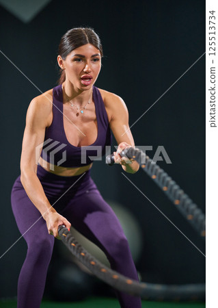 fit woman in purple activewear uses battle ropes enhance strength and endurance in gym setting. intense workout showcases her dedication and effort in dynamic environment. 125513734