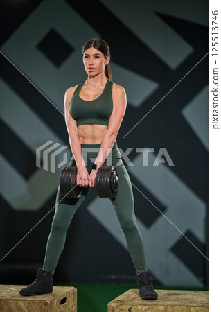 woman is engaged in strength training session gym while lifting dumbbell. She is wearing green activewear and is focused on her workout routine demonstrating dedication. 125513746