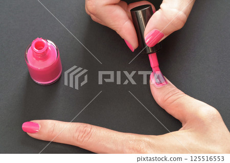 Applying pink nail polish - closeup photo of woman hands, little brush and fingernails on gray / black background Applying pink nail polish - closeup photo of woman hands, little brush and fingernails on gray / black background 125516553