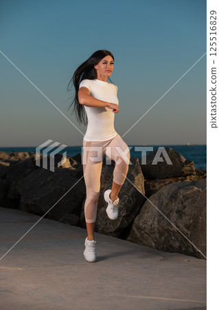 woman jogs along waterfront path showcasing her fitness and energy in early morning light. calm water and rocky shoreline create serene backdrop for her exercise. 125516829