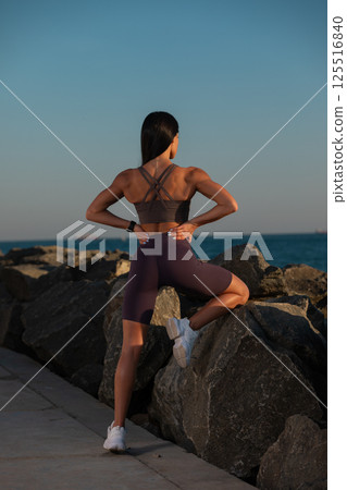 person in athletic wear performs stretch on large rocks water in coastal area during sunset. scenic view enhances wellness vibe. 125516840
