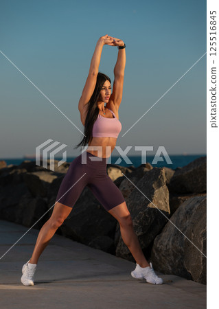 woman is stretching her arms overhead on coastal pathway lined rocks during sunset. She wears sports bra and shorts showcasing her commitment wellness and fitness. 125516845