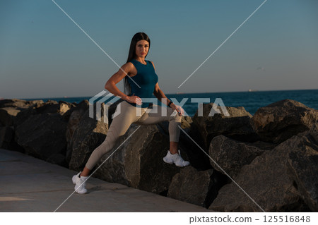 woman performs stretching exercise balancing on rocky shoreline as sun sets in background. ocean waves create serene atmosphere while she focuses on her workout. woman performs stretching exercise balancing on rocky shoreline as sun sets in background. ocean waves create serene atmosphere while she focuses on her workout. 125516848