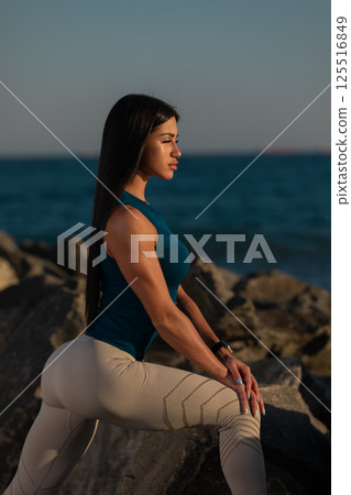 woman engaged in yoga pose on rocky terrain beside ocean sunset. She displays focus and strength her long hair flowing gently in breeze. 125516849