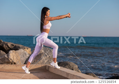 woman is stretching water's edge on sunny day. She wears light athletic clothing and focuses on her fitness routine while enjoying ocean view and tranquility. woman is stretching water's edge on sunny day. She wears light athletic clothing and focuses on her fitness routine while enjoying ocean view and tranquility. 125516854