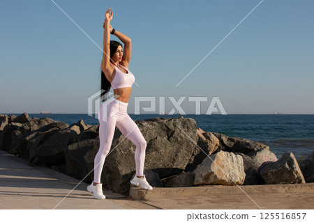 person engages in stretching exercise along rock lined beach. sun sets over water casting warm glow on fitness enthusiast dressed in light workout clothes. person engages in stretching exercise along rock lined beach. sun sets over water casting warm glow on fitness enthusiast dressed in light workout clothes. 125516857
