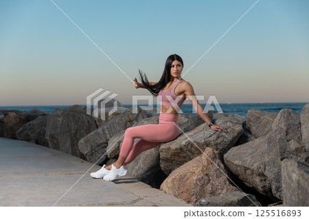 fitness enthusiast sits on large rocks water enjoying sunset. She wears athletic clothing showcasing strength and relaxation shoreline. 125516893