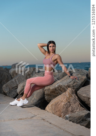 wellness advocate relaxes on large rocks ocean showcasing fitness attire against backdrop of colorful sunset. scene conveys tranquility and health. 125516894