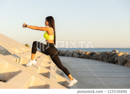 woman in bright top engages in lunges on outdoor structure near water enjoying sunset while focusing on her fitness routine and overall wellness. 125516898