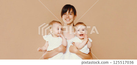 Portrait of happy smiling young mother holding twin babies on brown background Portrait of happy smiling young mother holding twin babies on brown background 125517036