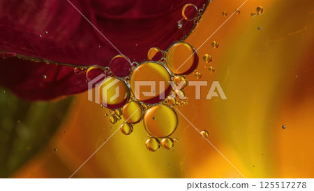 Intricate droplets of liquid hover delicately beneath a vibrant rose petal creating a mesmerizing dance of colors. The warm background adds depth and allure to this captivating moment. 125517278