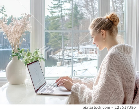 A woman typing on a laptop at a bright workspace with natural light, a vase of white flowers in the background, productivity and home office concepts 125517380