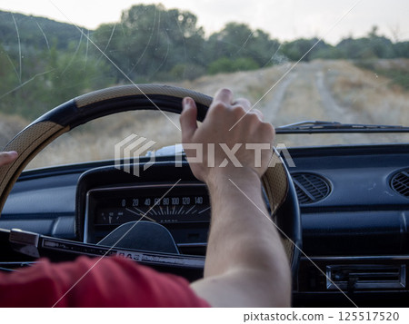 A person is gripping the steering wheel of an old car while navigating a dirt road surrounded by lush green trees and gentle hills during the late afternoon. A person is gripping the steering wheel of an old car while navigating a dirt road surrounded by lush green trees and gentle hills during the late afternoon. 125517520