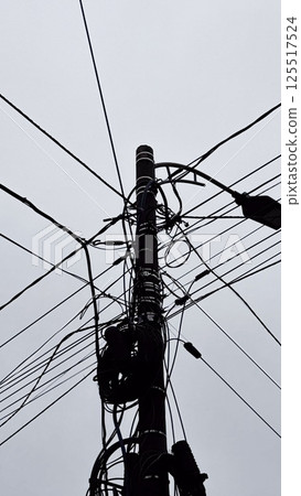 A tall power pole stands prominently amid a gray sky, with numerous wires crisscrossing overhead, demonstrating the complexity of urban electrical infrastructure during a cloudy day. A tall power pole stands prominently amid a gray sky, with numerous wires crisscrossing overhead, demonstrating the complexity of urban electrical infrastructure during a cloudy day. 125517524