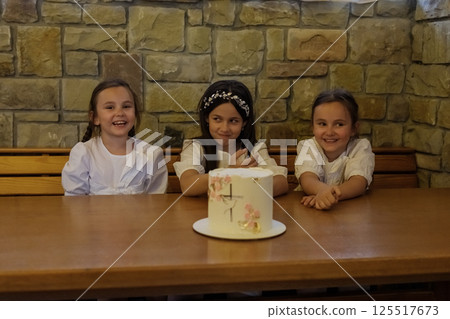 The girls in matching blouses and a dress form a charming group around the cake. The twin girls with two braids look fascinated. 125517673