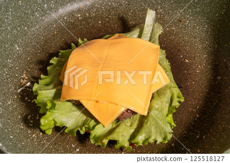 A juicy burger patty topped with melted cheese rests on lettuce inside a frying pan, showcasing the burger cooking process in progress. 125518127