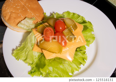 An open-faced burger with a sesame bun, lettuce, cheese, pickles, and cherry tomatoes on a white plate, highlighting vibrant burger preparation. An open-faced burger with a sesame bun, lettuce, cheese, pickles, and cherry tomatoes on a white plate, highlighting vibrant burger preparation. 125518131