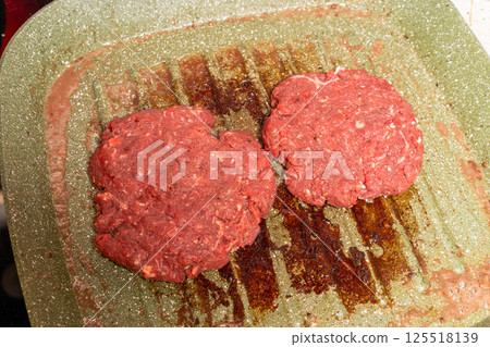Two raw burger patties on a speckled green grill pan, with grease marks emphasizing the homemade preparation process before grilling. 125518139