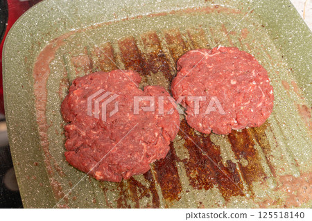 Two raw burger patties on a speckled green grill pan, with grease marks emphasizing the homemade preparation process before grilling. 125518140