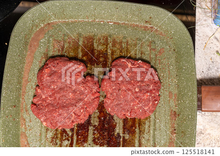 Two raw burger patties on a speckled green grill pan, with grease marks emphasizing the homemade preparation process before grilling. Two raw burger patties on a speckled green grill pan, with grease marks emphasizing the homemade preparation process before grilling. 125518141