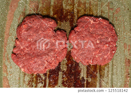 Two raw burger patties on a speckled green grill pan, with grease marks emphasizing the homemade preparation process before grilling. 125518142